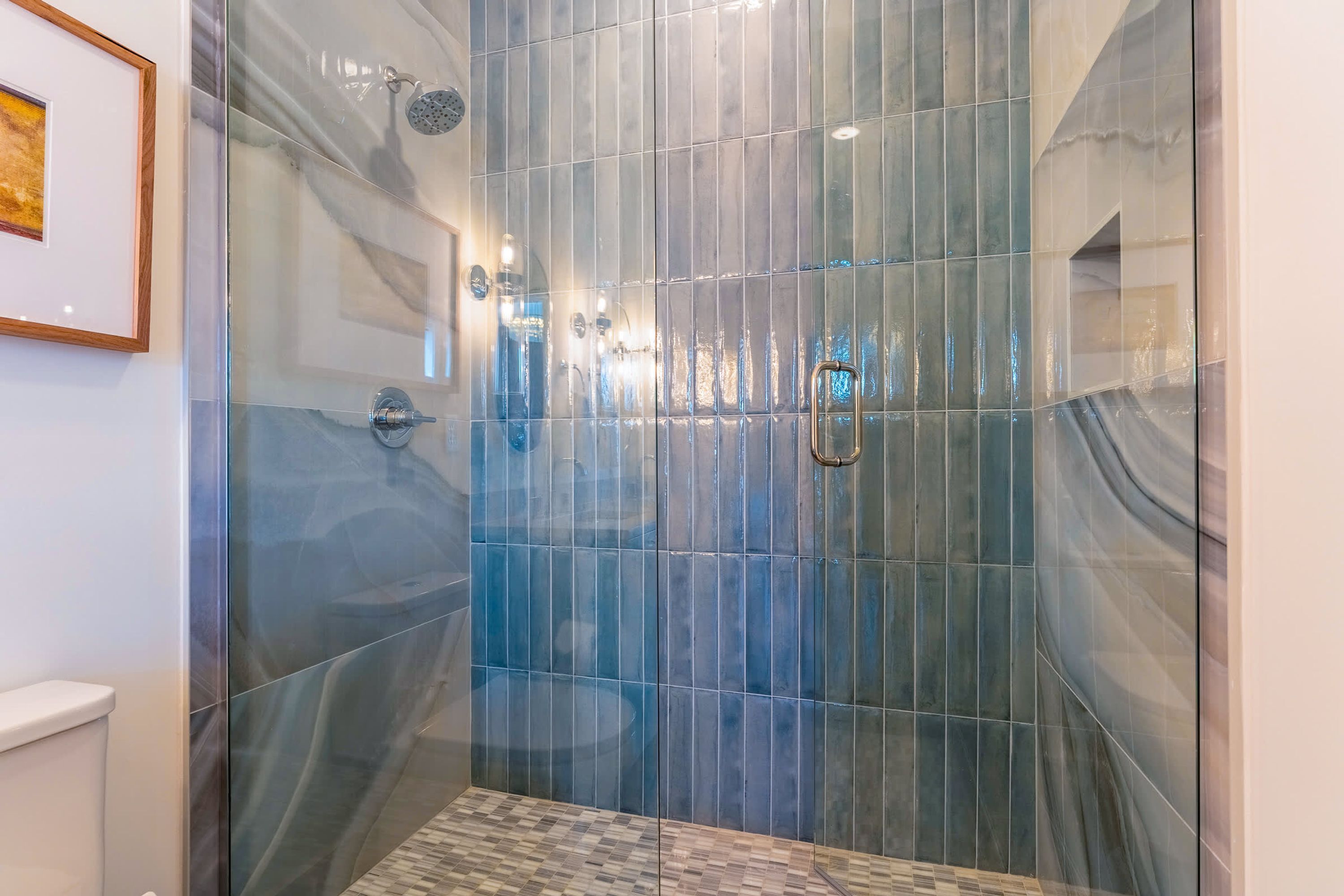 Une douche fermée par des parois en verre, avec un mur carrelé, une barre d’appui et un tableau encadré accroché au mur.