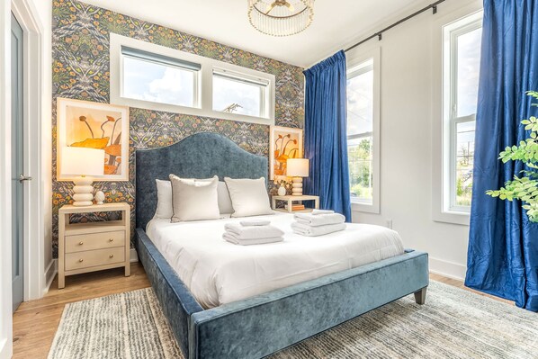 Ein Schlafzimmer mit einem großen Bett, zwei Nachttischen, einer Chandelüster und Blick ins Freie.