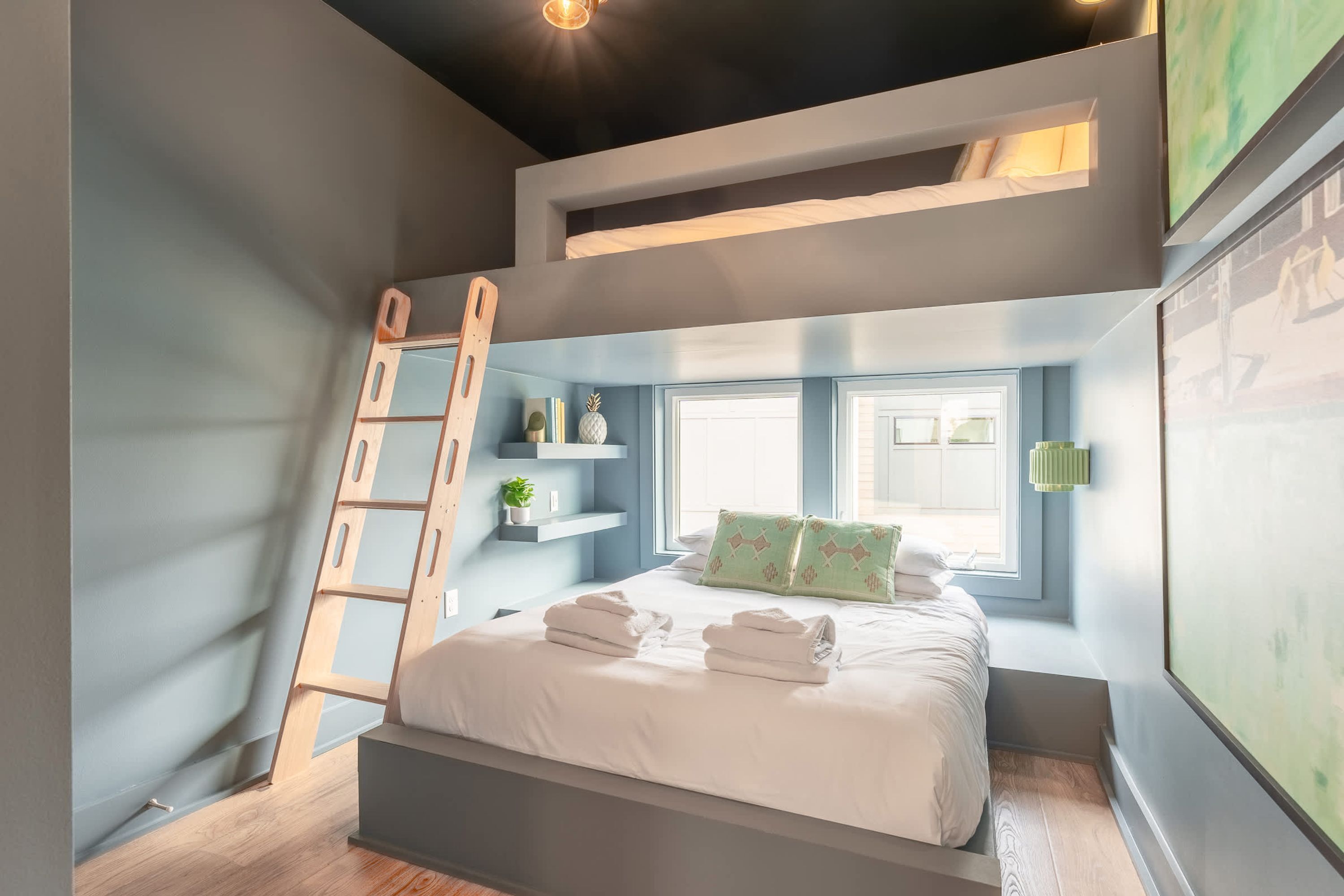 A bedroom with a bunk bed, a ladder, a window with daylight, and a green wall-mounted shelf with items.