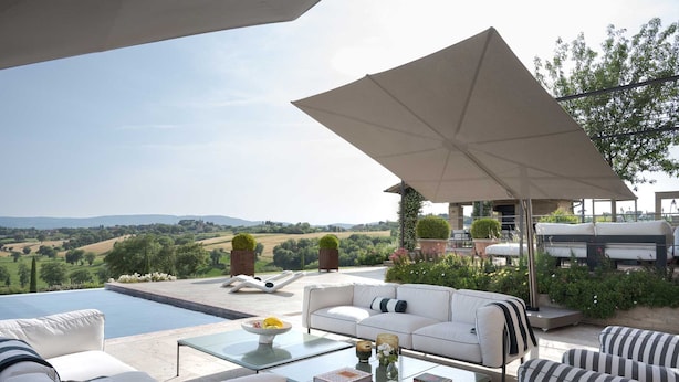 A luxurious outdoor seating area with a pool, white sofas, striped cushions, and a large umbrella providing shade. - Il Mandorlo by Boutique (San Quirico d'Orcia)