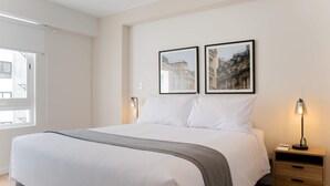 A neatly made bed with white linens and a gray throw blanket, two framed pictures on the wall, a bedside lamp, and a wooden nightstand with a drawer. - w Comfortable 2BR Stay in Miraflores (Lima)