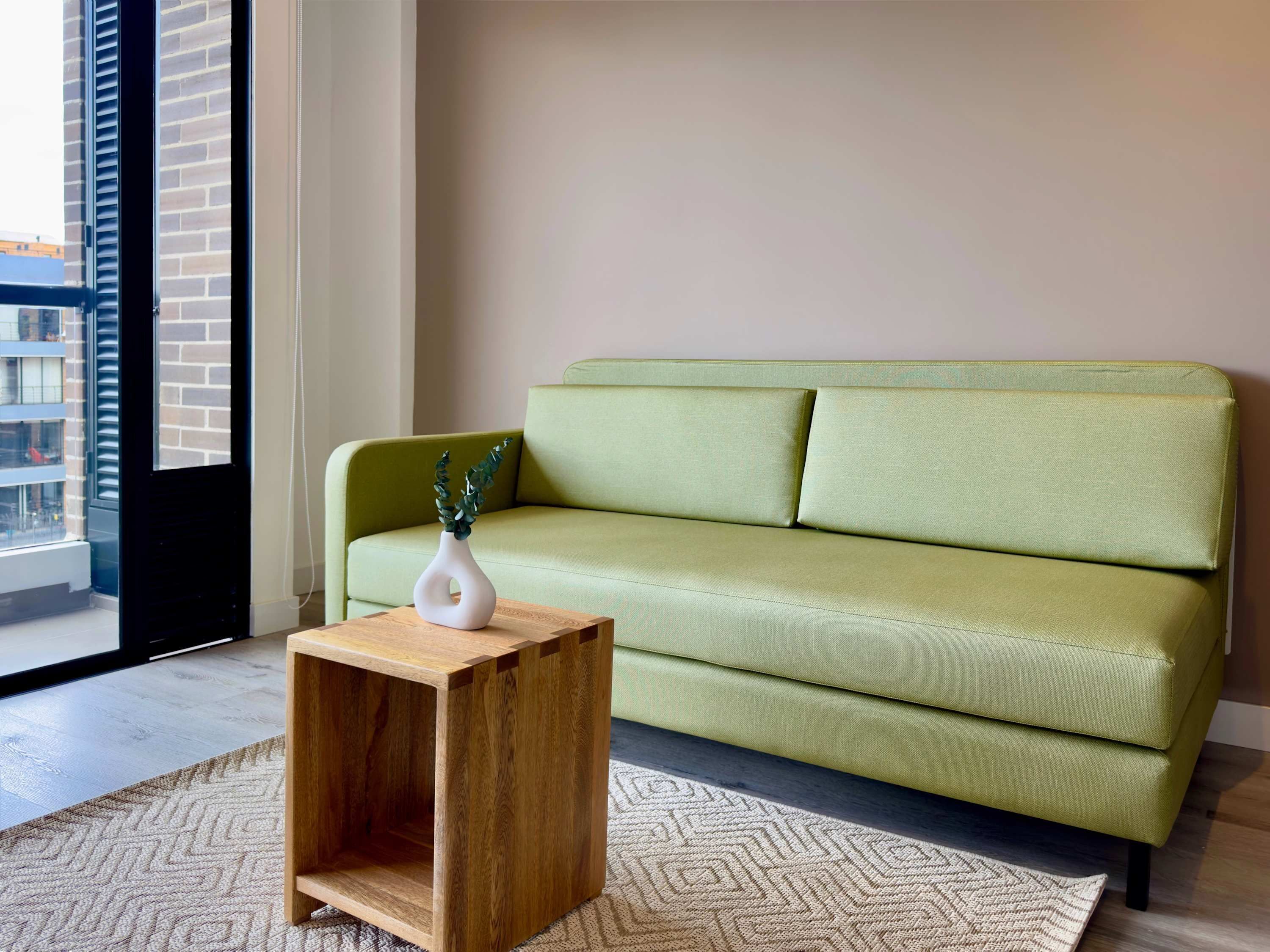 Un sofá verde con un jarrón blanco y una mesita de madera en una habitación con alfombra estampada y vistas a edificios.