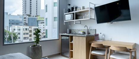 A modern kitchen with a built-in refrigerator, microwave, and a flat-screen TV. There is a dining area with a wooden table and chairs, and a large window offering a city view.