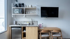 Cozinha compacta com geladeira embutida, micro-ondas e cafeteira na bancada. Há uma mesa de jantar de madeira com duas cadeiras e uma TV fixada na parede.