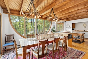 Un intérieur en bois comprenant une table à manger, des chaises, un lustre et de grandes fenêtres donnant sur la forêt.