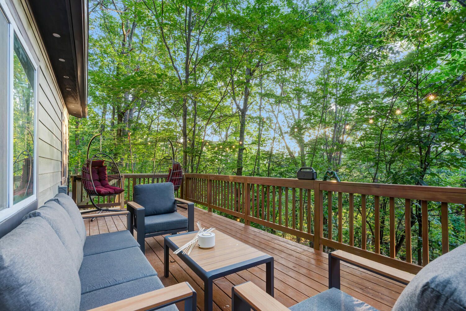 Terraza de madera con mobiliario exterior, incluyendo un sofá y sillas, con vistas a una zona boscosa.