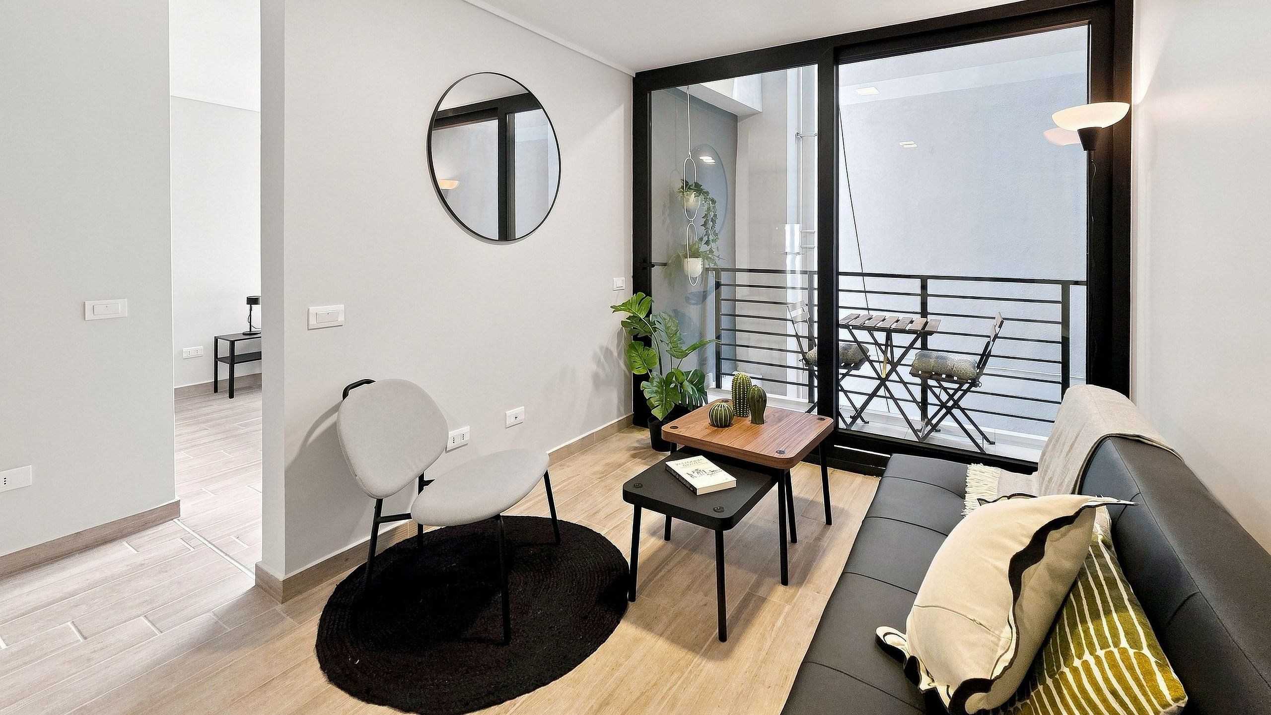A modern living room with a black sofa, a round mirror, a small table with a book, and a balcony with a railing.