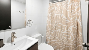 Un baño con lavamanos blanco, inodoro, espejo y cortina de ducha estampada con hojas.