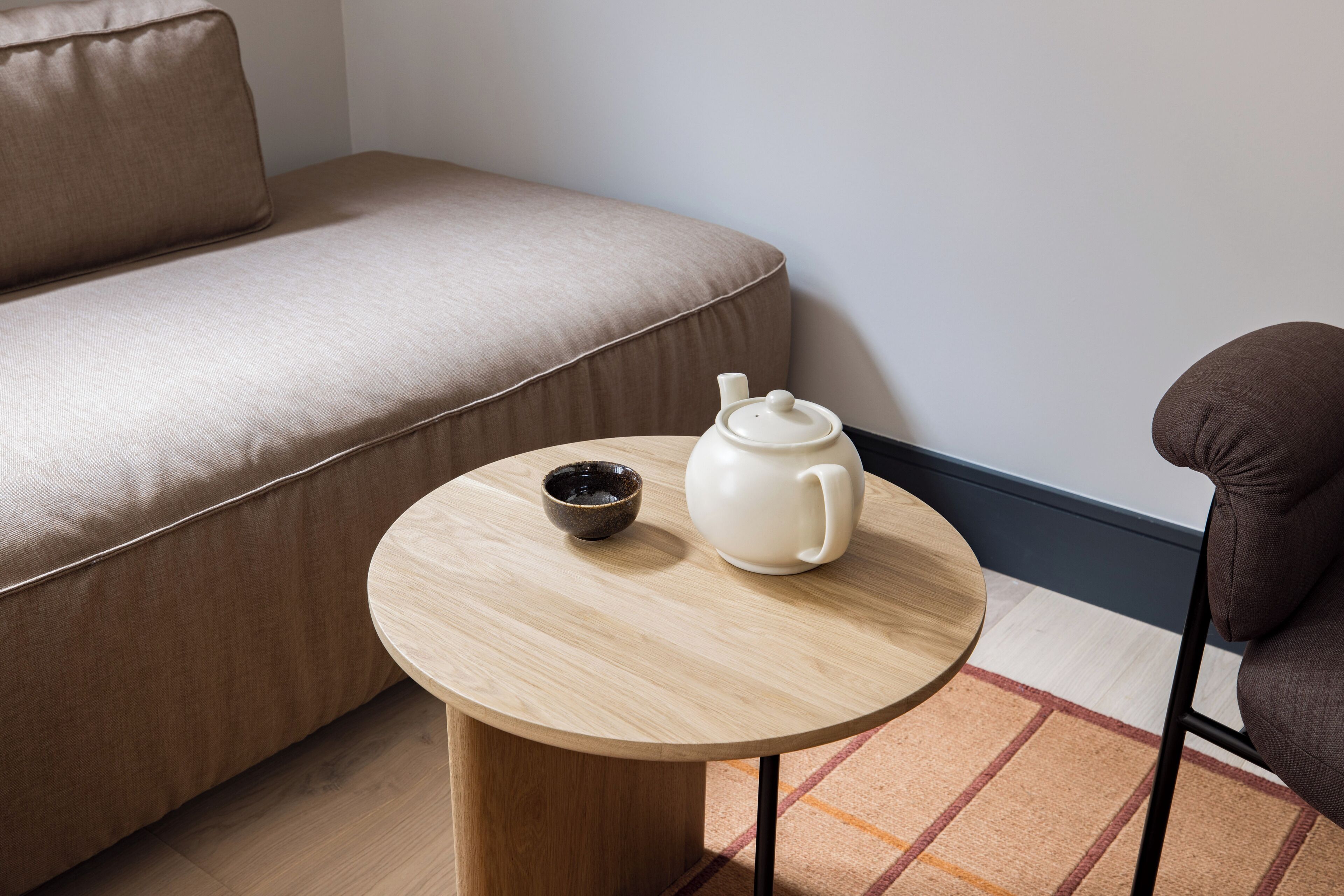 A round wooden table with a white teapot and a dark bowl on a beige sofa.