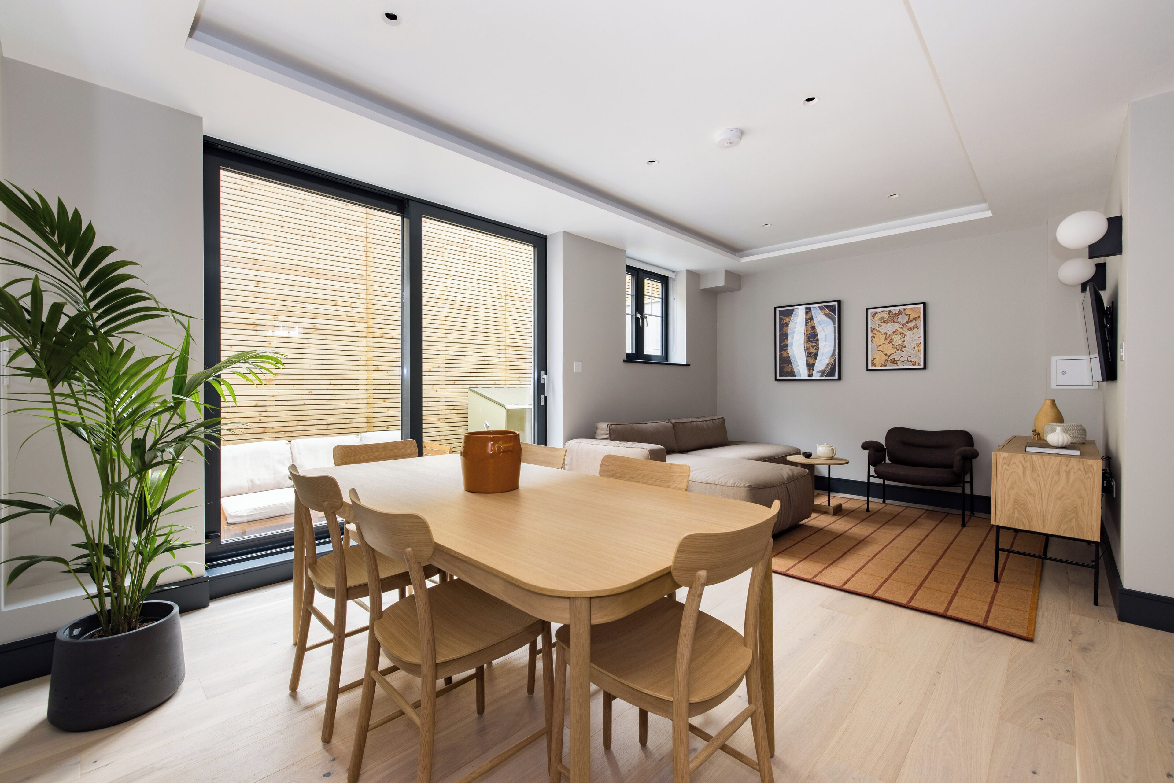 Un salon moderne avec une table à manger en bois, des chaises, un canapé et une table basse. Une grande fenêtre avec des stores, une plante en pot et des œuvres d’art ornent le mur.