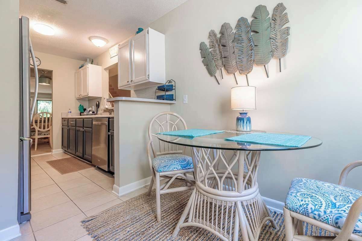A modern kitchen with white cabinets, a dining area with a glass table and wicker chairs, and a decorative leaf wall piece.