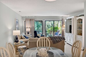 Una sala de estar con una mesa de vidrio, un sofá, un mueble para televisión y vistas a la vegetación exterior.