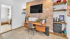 Une chambre moderne avec un mur d’accent en briques, un téléviseur à écran plat, un bureau en bois, une chaise, un petit réfrigérateur et des étagères contenant divers objets.