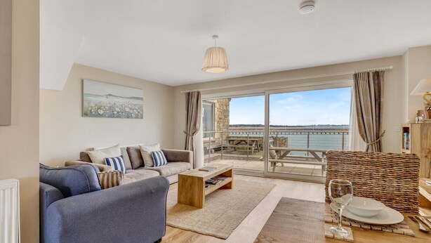 Un salon moderne avec un canapé, une table basse et une table à manger pour deux personnes. Une grande fenêtre offre une vue sur la mer et un quai.