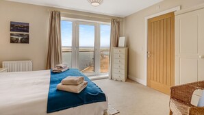 Une chambre à coucher comprenant un lit, une commode, une chaise et une vue sur la plage, accessible par une porte coulissante.