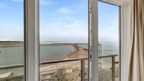 Une vue depuis une fenêtre donnant sur une zone côtière, avec une jetée s’avançant dans l’eau, une plage de sable et un promontoire rocheux.