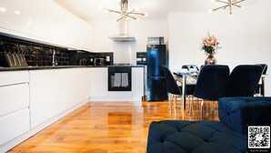 Una cocina moderna con armarios blancos, encimera negra y horno empotrado. Hay un comedor con mesa y sillas, y un sofá negro en primer plano.