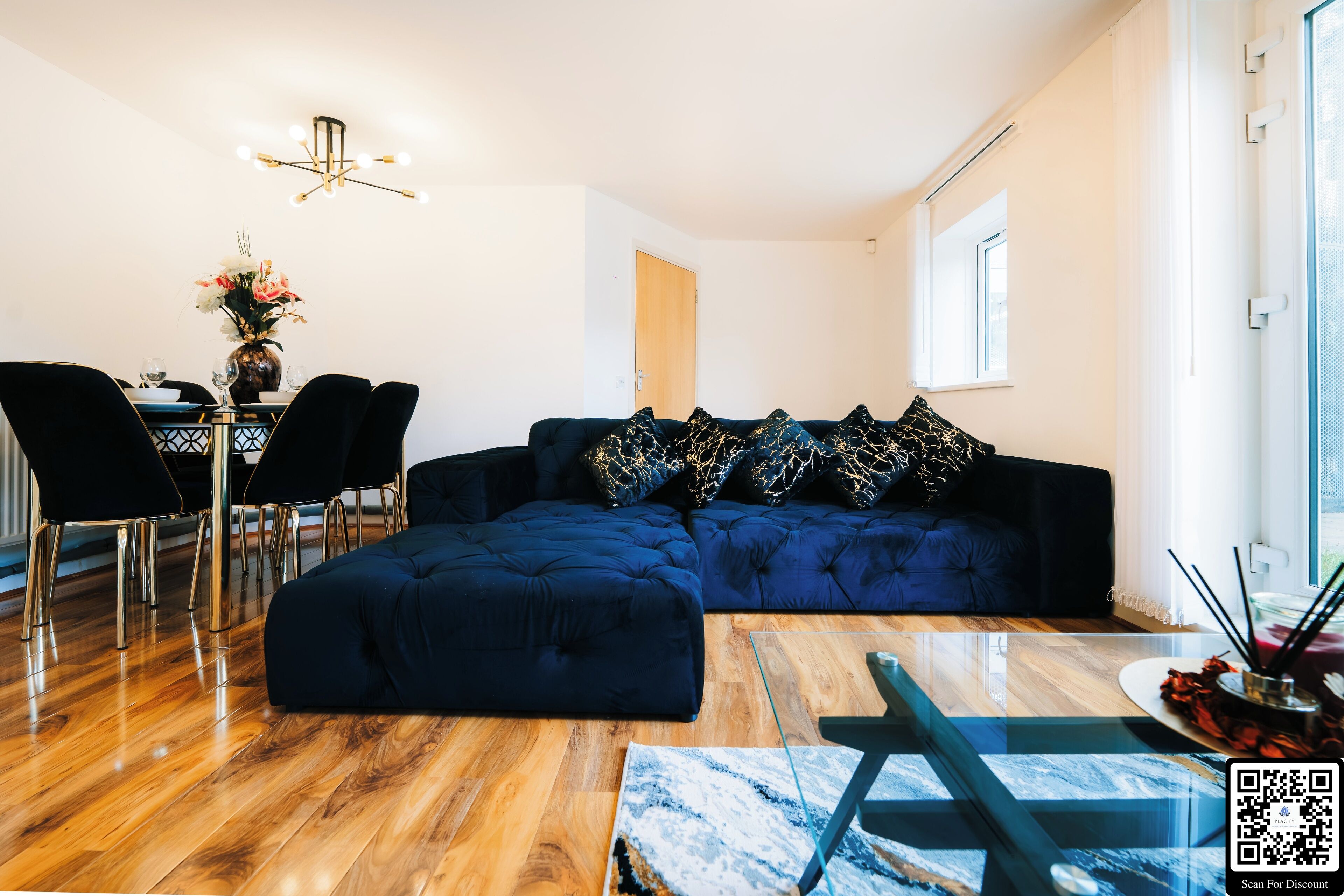 Een moderne woonkamer met een donkerblauwe modulaire bank, een eethoek met een glazen tafel en een kroonluchter.