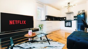 Una sala moderna con un televisor de pantalla plana grande que muestra 'NETFLIX', una mesa de centro de vidrio, un sofá azul oscuro y una zona de cocina con armarios blancos y una encimera negra.