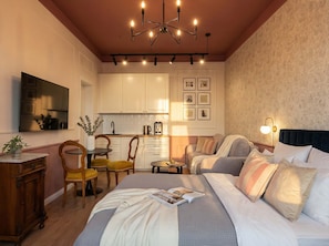 Een slaapkamer met een bed, nachtkastje en een klein tafeltje. Er is een raam met gordijnen, een tv aan de muur en een klein tafeltje met stoelen.