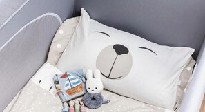 Ein Babybett mit weißem Kopfkissen, einem Bärengesicht-Design, einem kleinen Stofftier und einer getupften Decke.