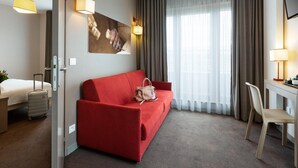 Ein Hotelzimmer mit rotem Sofa, Schreibtisch, Stuhl, Bett, Koffer und einem Bild an der Wand.