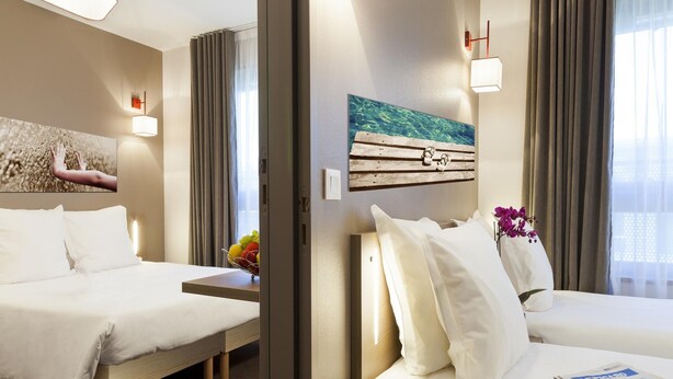 Ein Hotelzimmer mit zwei Betten, einem Holzkopfteil, einem Wandbild, einem kleinen Tisch mit Blumenvase und einem fenster mit Vorhängen.