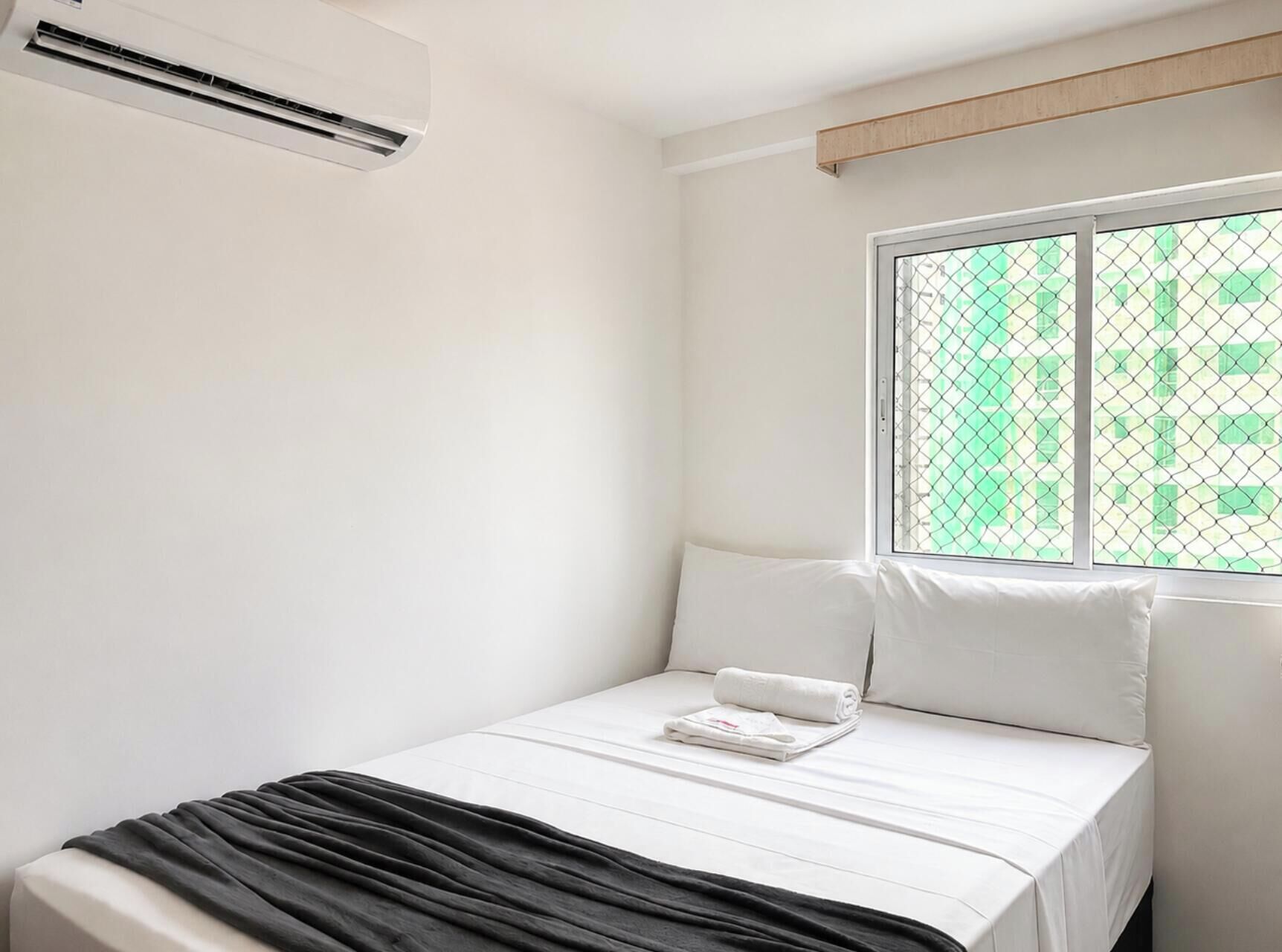 A hotel room with a bed, a window with a view of buildings, and an air conditioning unit.