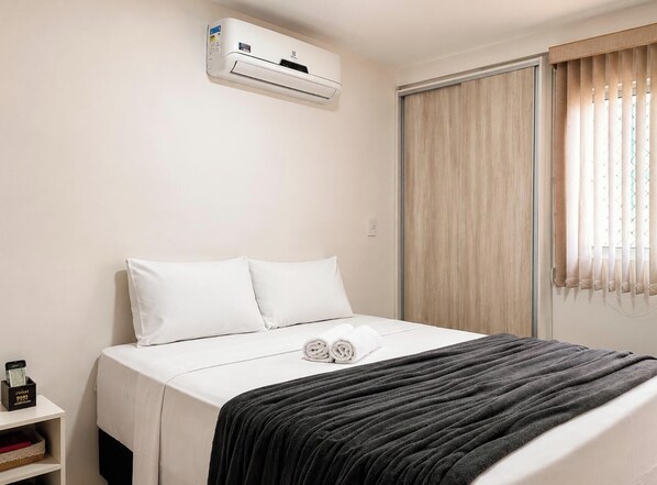 Ein Hotelzimmer mit einem Bett, einem Nachttisch mit Telefon und Ablagefach, einem Fenster mit Vorhängen und einer Klimaanlage an der Wand.