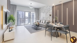 A modern living room with a sofa, dining table, and chairs, a TV mounted on the wall, and a large window with a city view. - Vogue | Downtown near Dubai Mall | Burj Khalifa (Dubai)