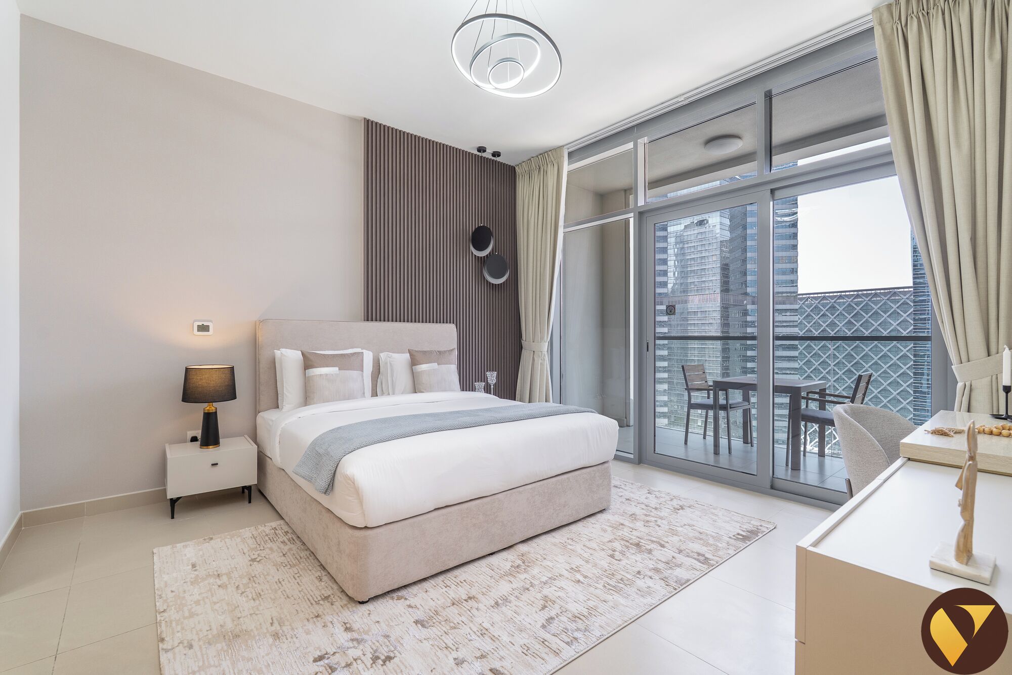 Une chambre moderne avec un grand lit, une table de chevet, une lampe, une vue sur la ville grâce à de grandes fenêtres et un balcon agrémenté de mobilier d’extérieur.