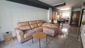 Una sala moderna con un sofá seccional color beige, una mesita de centro de madera y una cocina al fondo.