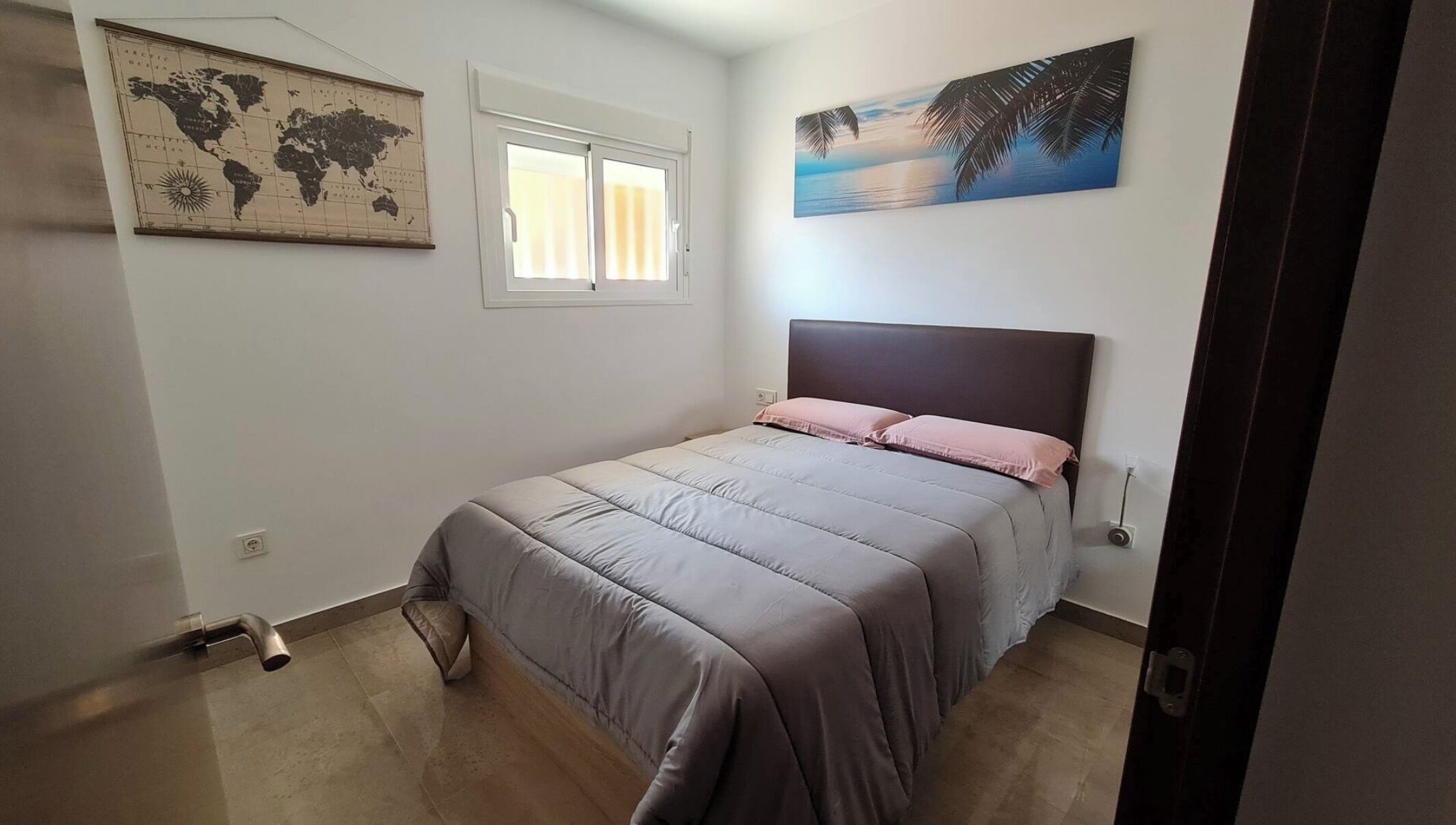 A bedroom with a bed, a world map, a window, and a painting of a sunset.