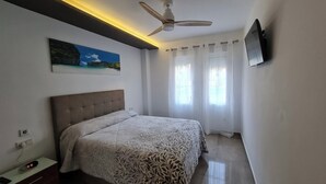 A bedroom with a bed, a ceiling fan, a painting, a TV, and a window with curtains.