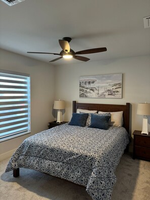 A bedroom with a bed, a ceiling fan, a window with blinds, a nightstand with a lamp, and a wall-mounted painting. - New- Villa white Peacock Exclusive Villa with all amenities, top location (Cape Coral)
