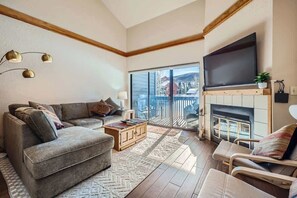 A living room with a sofa, armchair, fireplace, TV, and a view through a glass door.