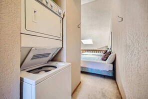 A laundry area with a front-loading washer and dryer, a bed with a mattress and pillows, and a small window above the bed.