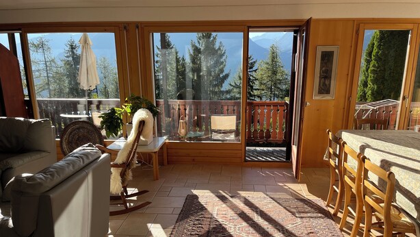 Ein geräumiges Wohnzimmer mit einer großen Schiebeglaswand, die auf einen Balkon führt, einem Essbereich mit Holztischen und einem Teppich mit Muster.