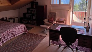 Ein gemütliches Zimmer mit schrägem Dach, einem Bett mit gemusterten Betttextilien, einem Schreibtisch mit Stuhl, einem Bücherregal und Bergblick durch ein Fenster.