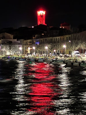 Une vue nocturne d’un front de mer avec des bateaux, une tour illuminée en rouge et des bâtiments le long de la côte.