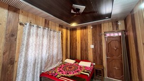 A bedroom with wooden walls, a ceiling fan, a bed with a patterned bedspread, and a wooden door. - Peace Forest Retreat (Chamoli)