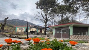 A building with a green roof and a fenced area with orange flowers in the foreground. - Peace Forest Retreat (Chamoli)
