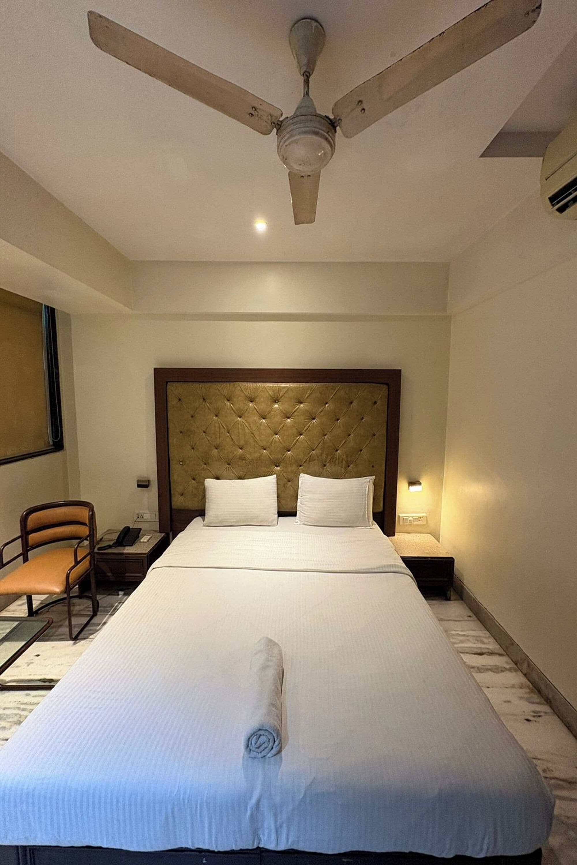 Una habitación de hotel con una cama grande, un ventilador de techo, un escritorio con una silla y una mesita de noche con lámpara.
