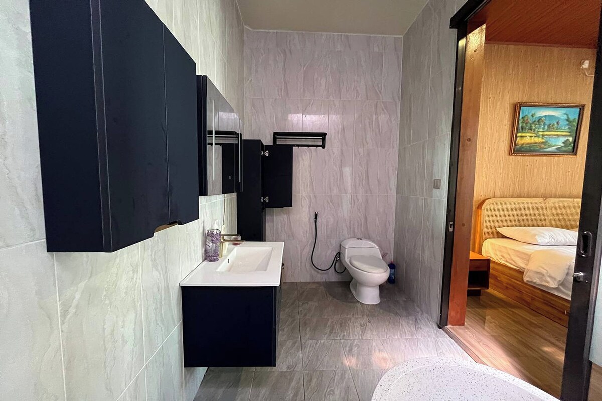 A modern bathroom with a white sink, black cabinet, and a toilet. There is a wooden door leading to a bedroom with a bed and a painting on the wall.