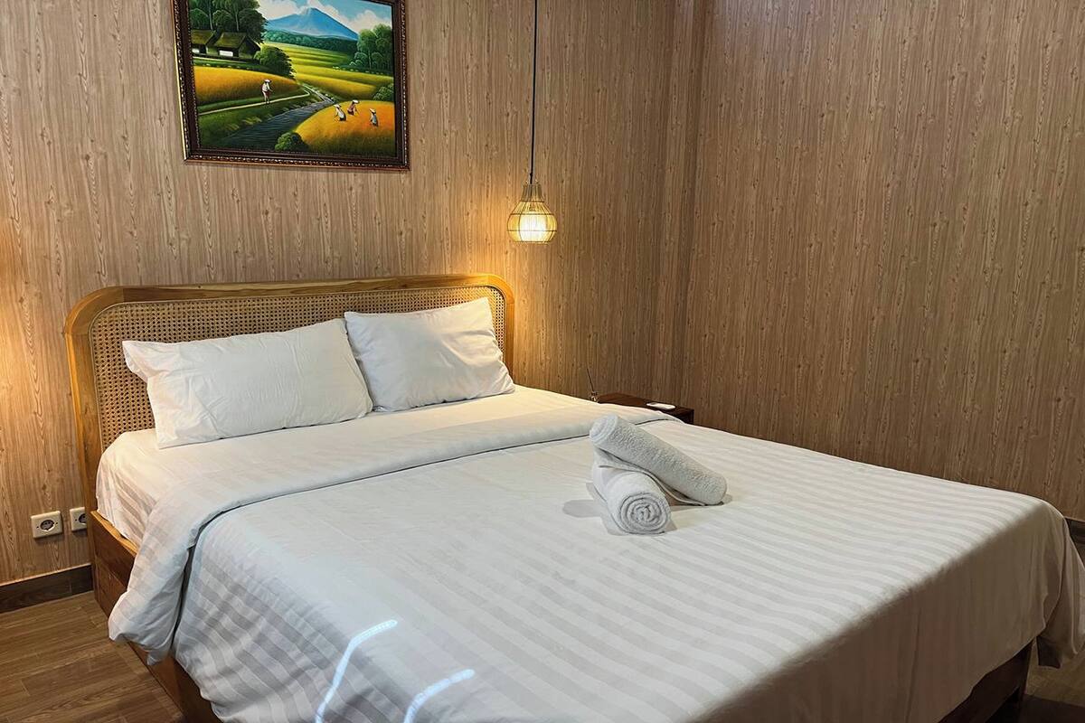A neatly made bed with white linens and a rolled towel, a wooden headboard, and a painting of a landscape on the wall.