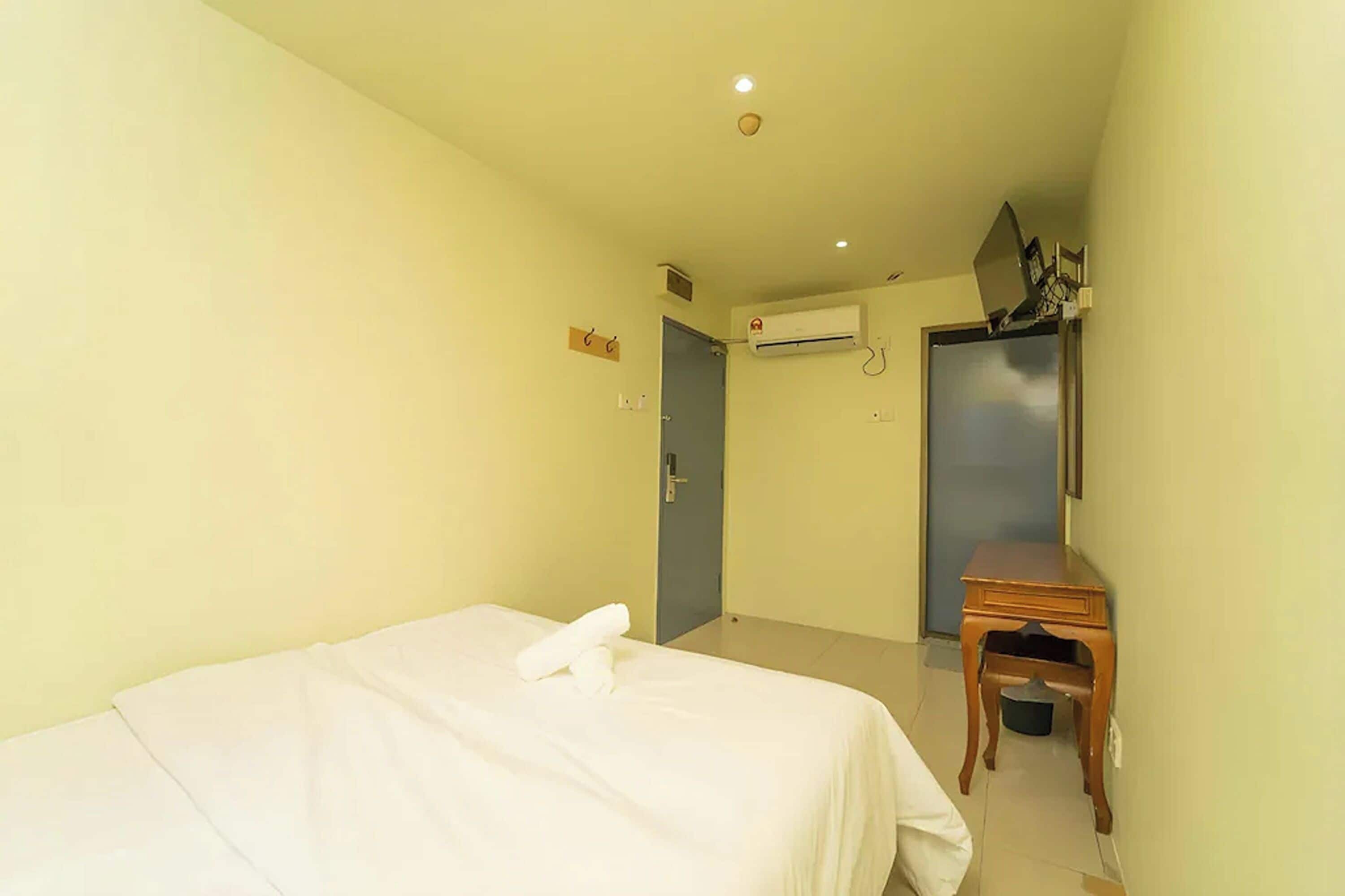 A hotel room with a bed, a small wooden table, an air conditioning unit, and a door.