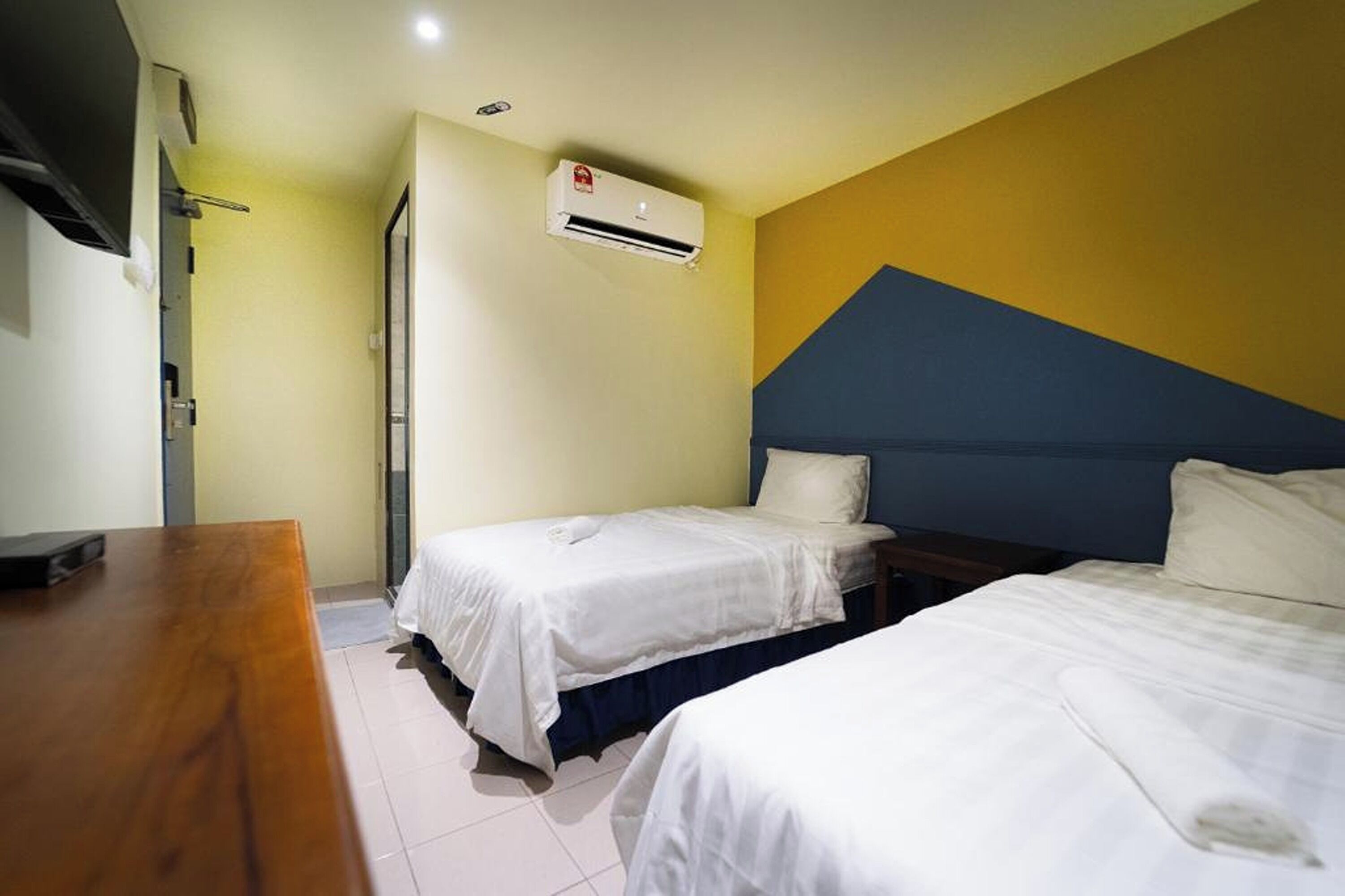 A hotel room with two beds, a wooden desk, an air conditioning unit, and a door.