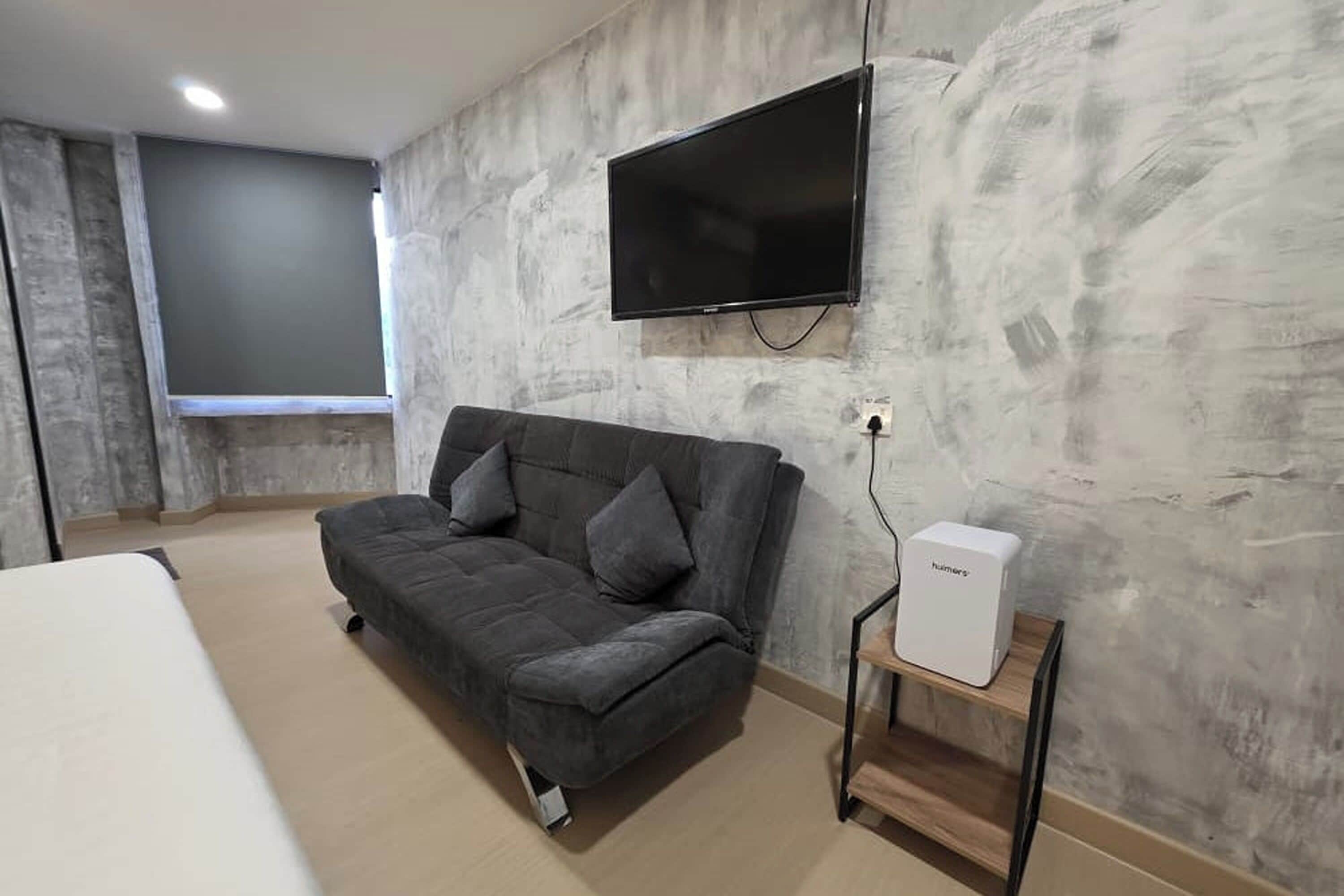 A modern room with a grey sofa, a flat-screen TV mounted on the wall, and a small white device on a wooden stand.