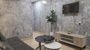 Un salon moderne avec un canapé gris, une petite table ronde agrémentée d’une plante, un téléviseur fixé au mur et un meuble TV en bois.