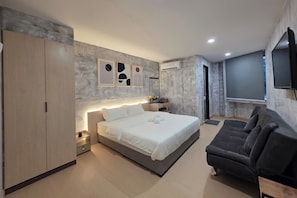 A modern bedroom with a bed, a dark sofa, a wardrobe, a nightstand, and a wall-mounted TV. - The Cozy Hotel (Sungai Petani)